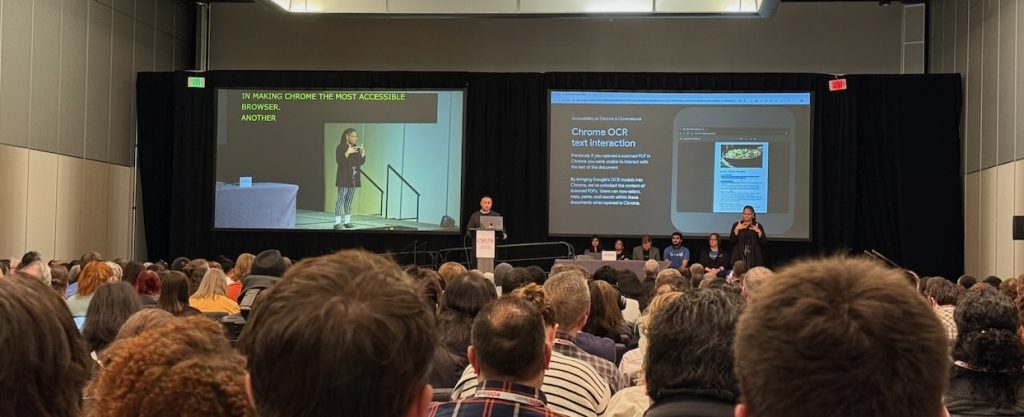 Crowd of heads with the presenter and their presentation at the far end of the hall.