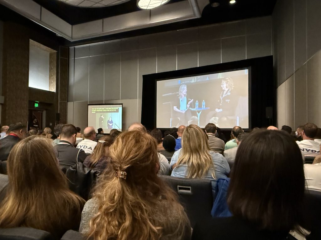 A crowd of people sitting down in front of a big screen showing two women talking, and a second screen with captions and sign language.